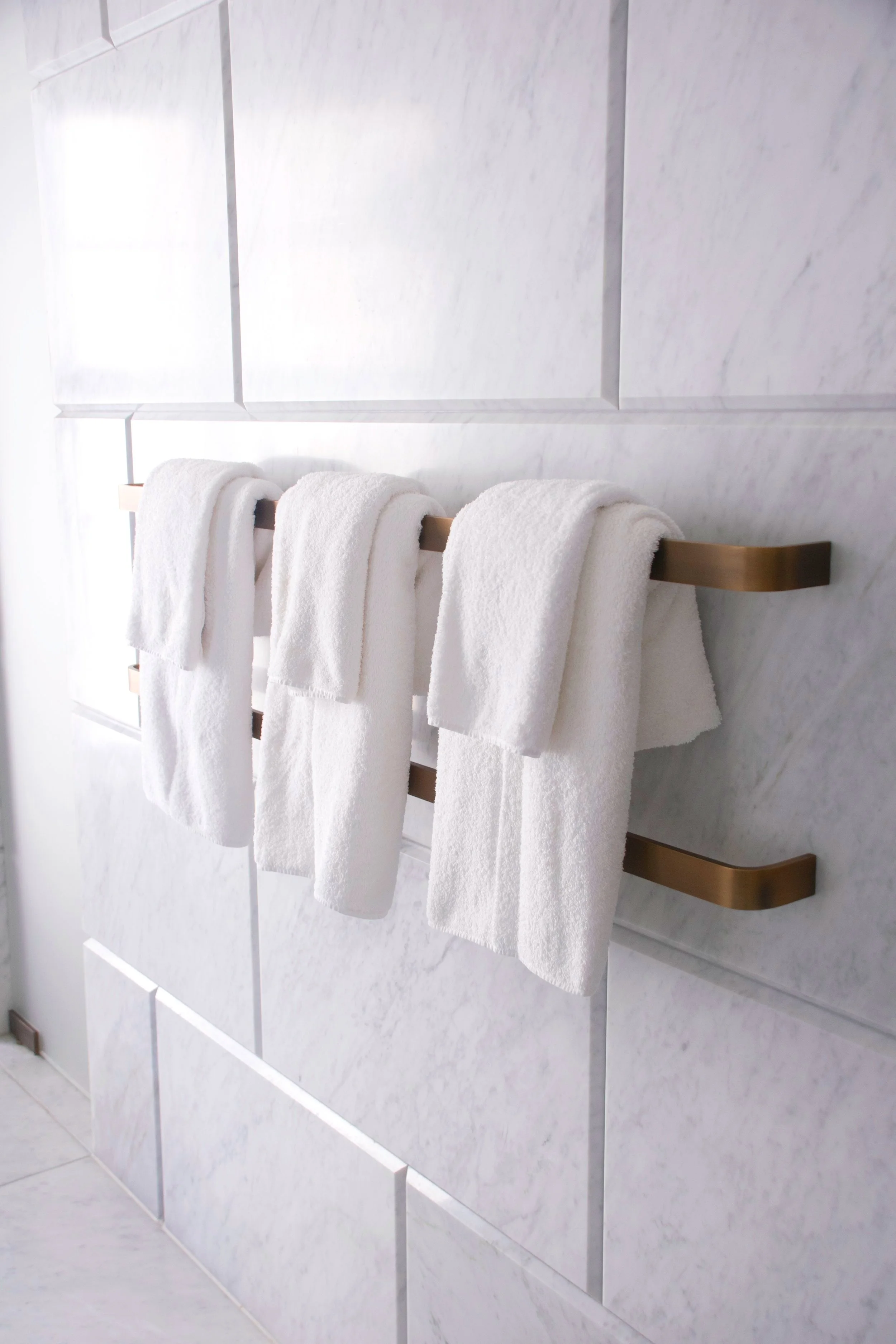 Three white towels hanging on a gold towel rack mounted on a marble wall in a bathroom.
