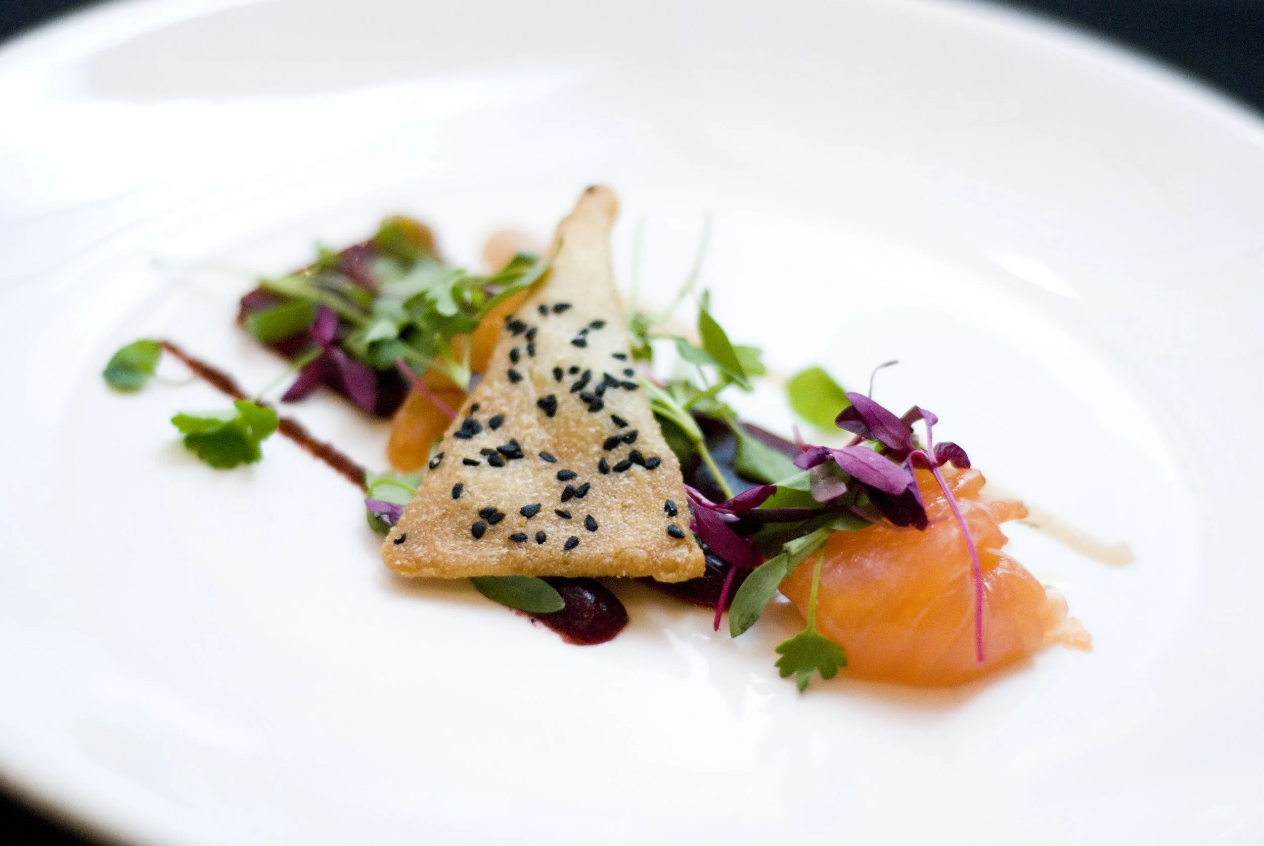 An elegant plated dish featuring a sesame seed-topped triangular cracker, microgreens, orange-colored sauce, and a garnish of microgreens and sauce drizzles on a white plate.