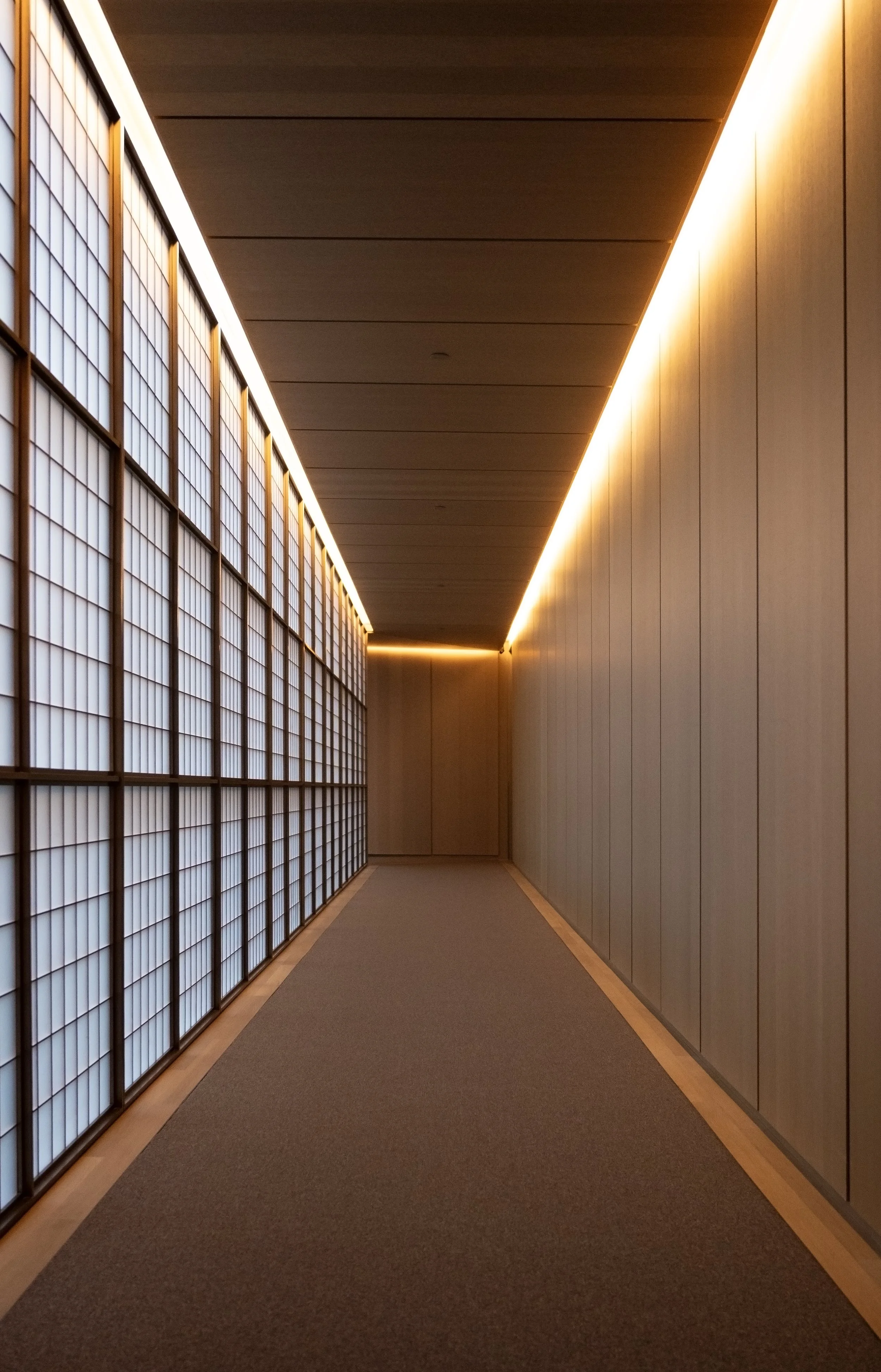 Empty corridor with wooden walls, ceiling, and floor edge, illuminated by warm indirect lighting along the ceiling and wall.
