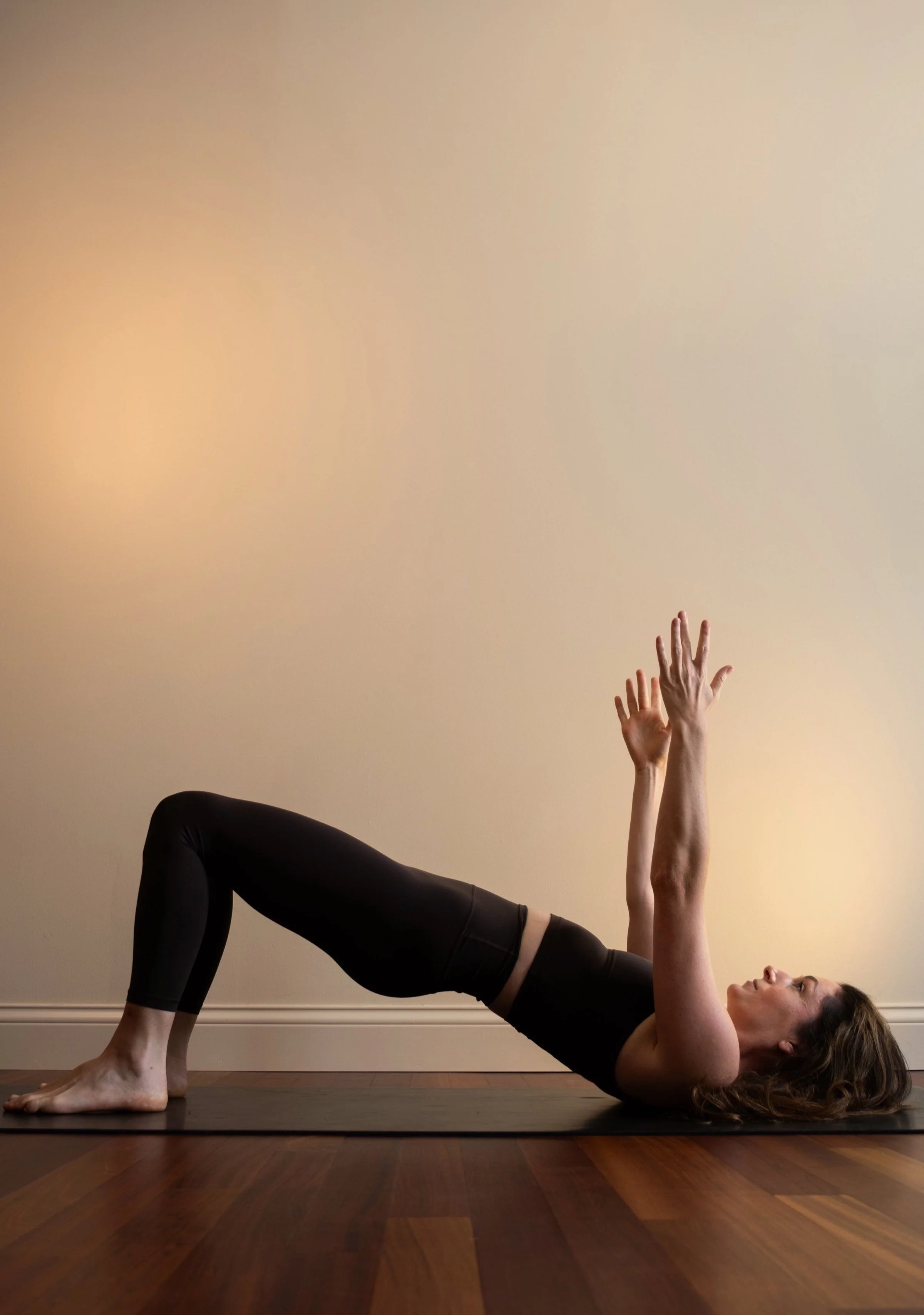 Woman practicing yoga indoors, lying on her back on a yoga mat with her knees bent, arms extended upward, looking up.