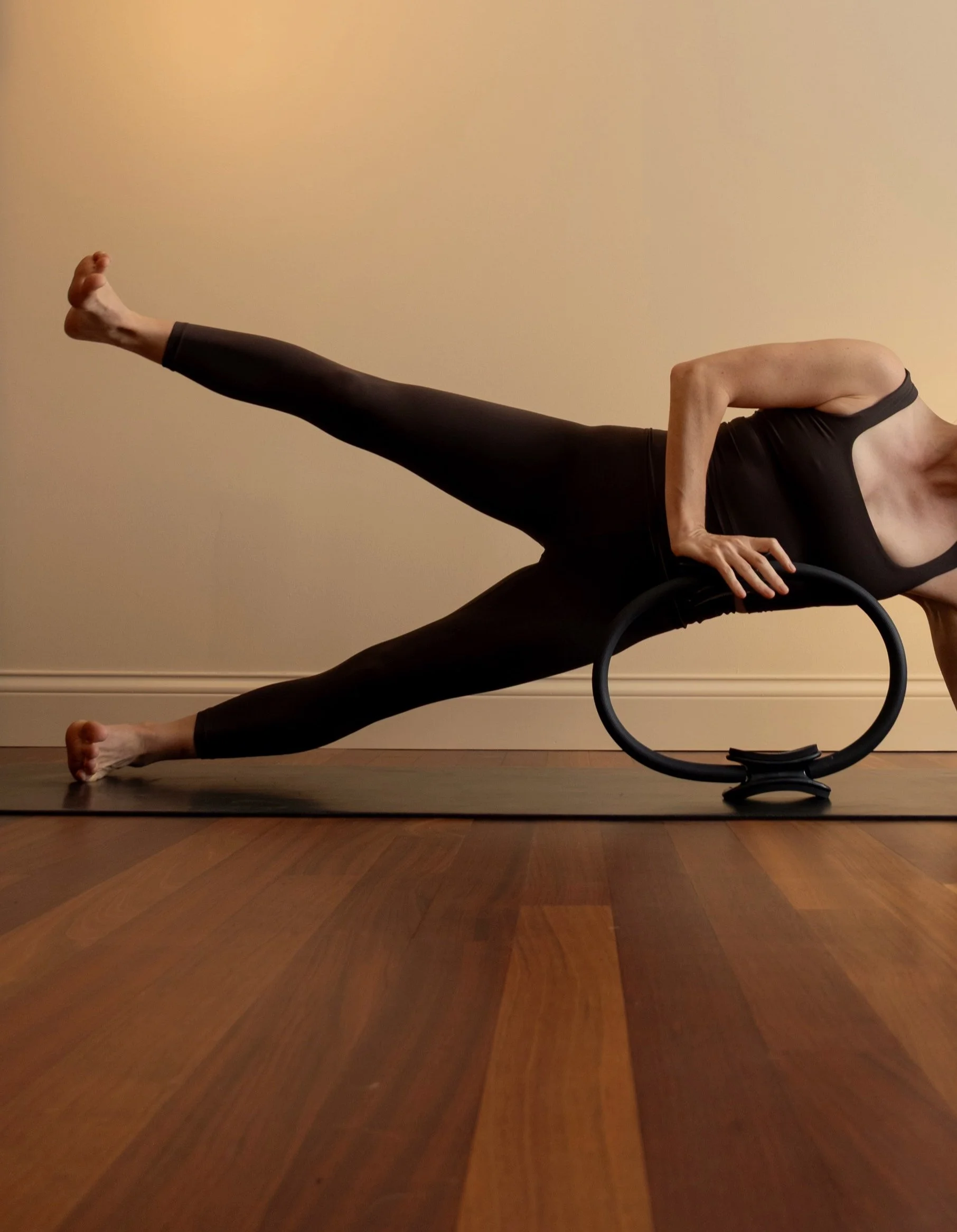 A person practicing yoga on a yoga mat, lying on their side, propped up on one arm, with one leg extended straight out and the other leg bent, holding a circular yoga prop.