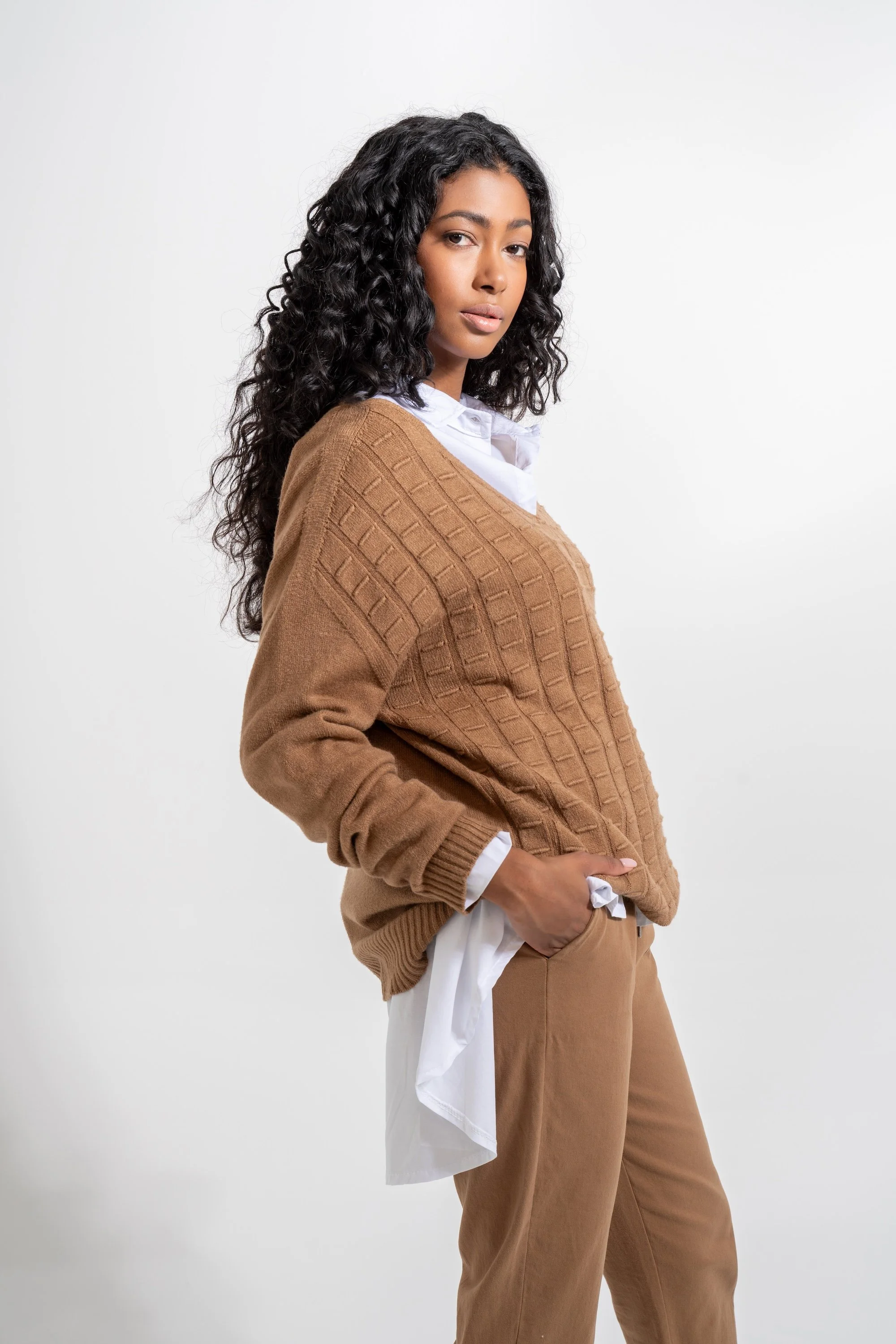 Young woman with curly hair wearing a brown sweater over a white shirt and brown pants, standing against a plain white background.