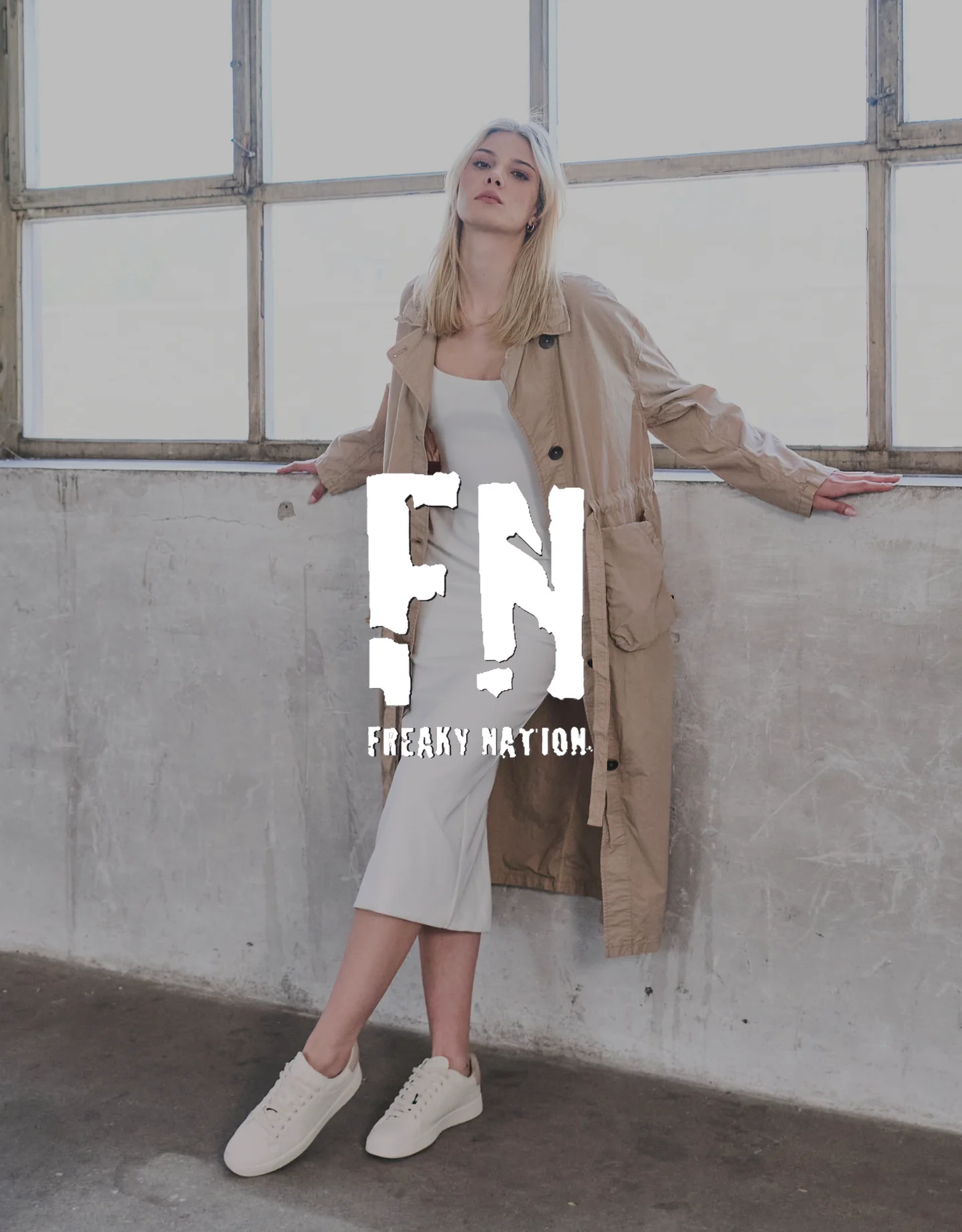 Young woman in casual beige and white clothing standing in an industrial-style room with large windows, with a logo overlaid that reads 'Freaky Nation'.