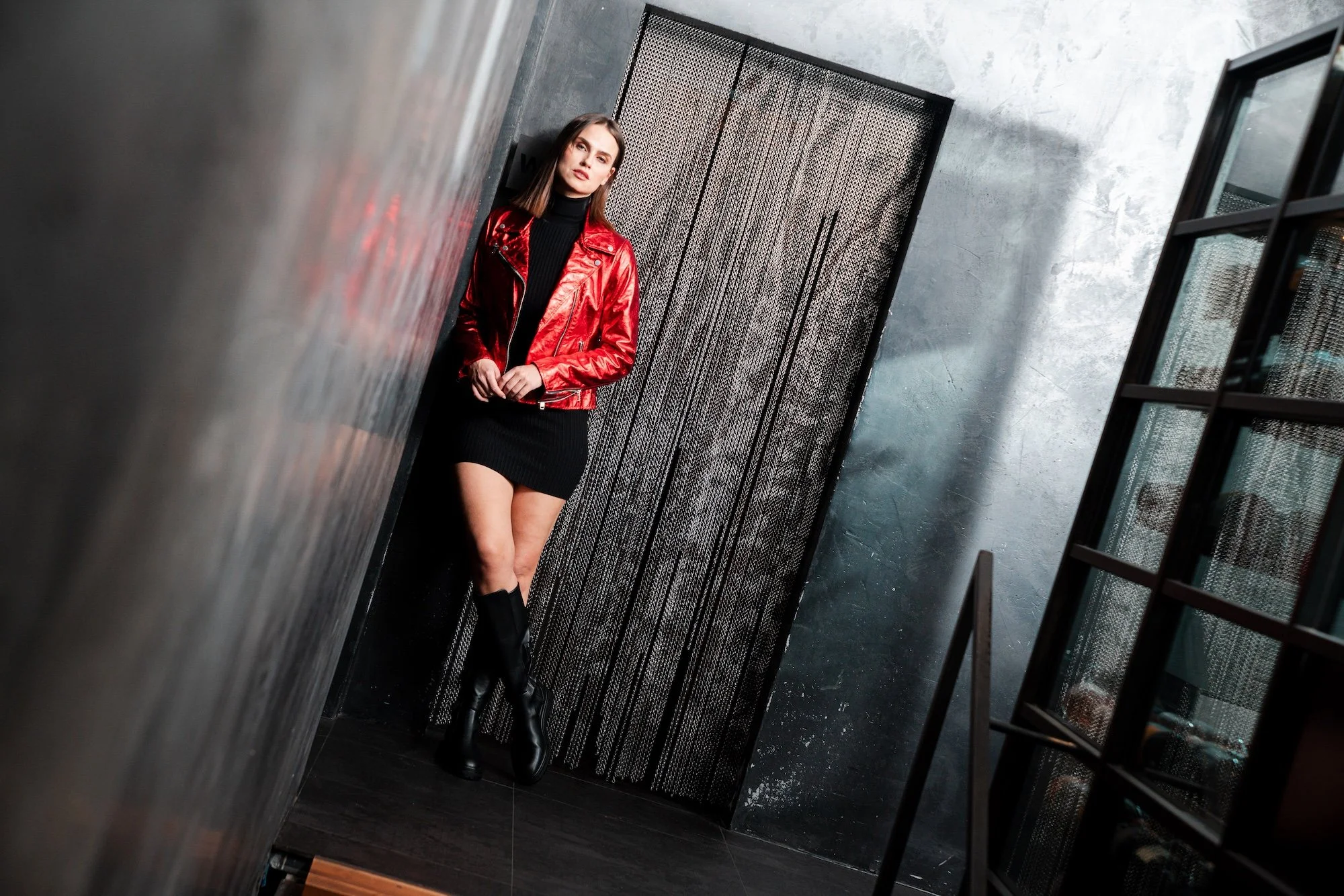A woman in a red leather jacket, black turtleneck, and black boots leaning against a wall in a modern industrial-style space with gray walls and black metal accents.