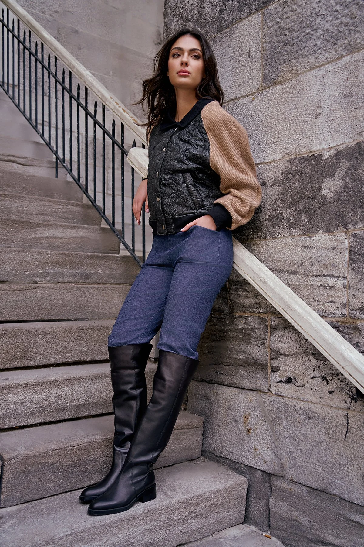 A woman standing on stone stairs next to a stone wall, wearing a beige and black varsity jacket, blue pants, and black knee-high boots.