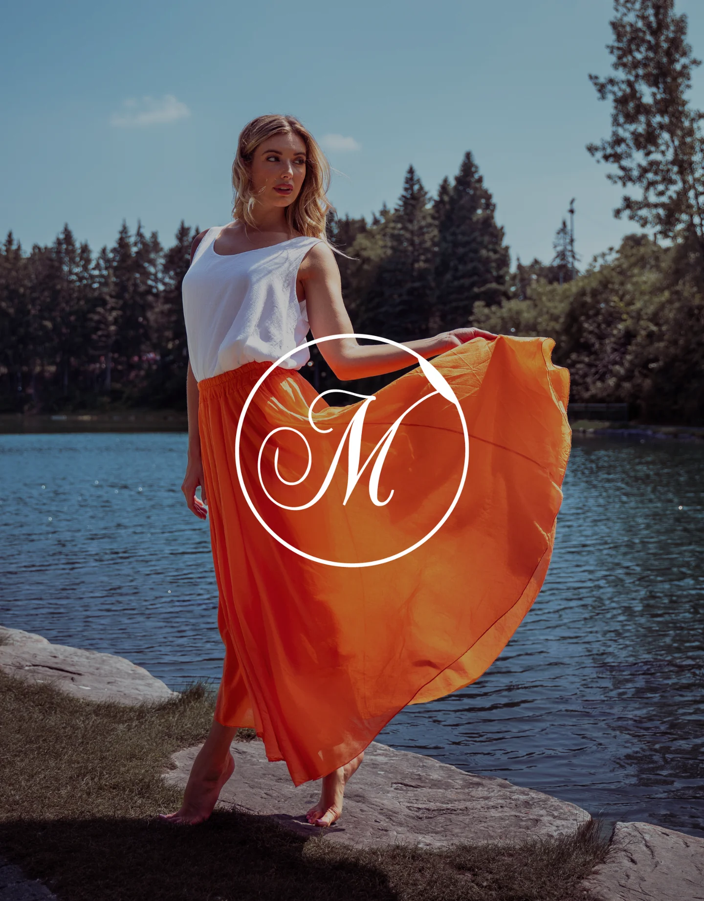 A woman wearing a white sleeveless top and an orange maxi skirt standing by a river with trees in the background.