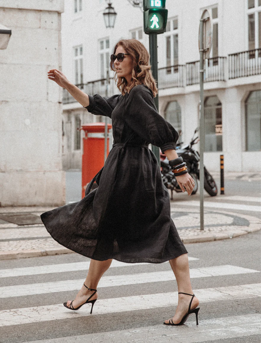 A woman crossing the street at a crosswalk while wearing a black dress and high heels, with sunglasses and bracelets, during daytime in an urban area.
