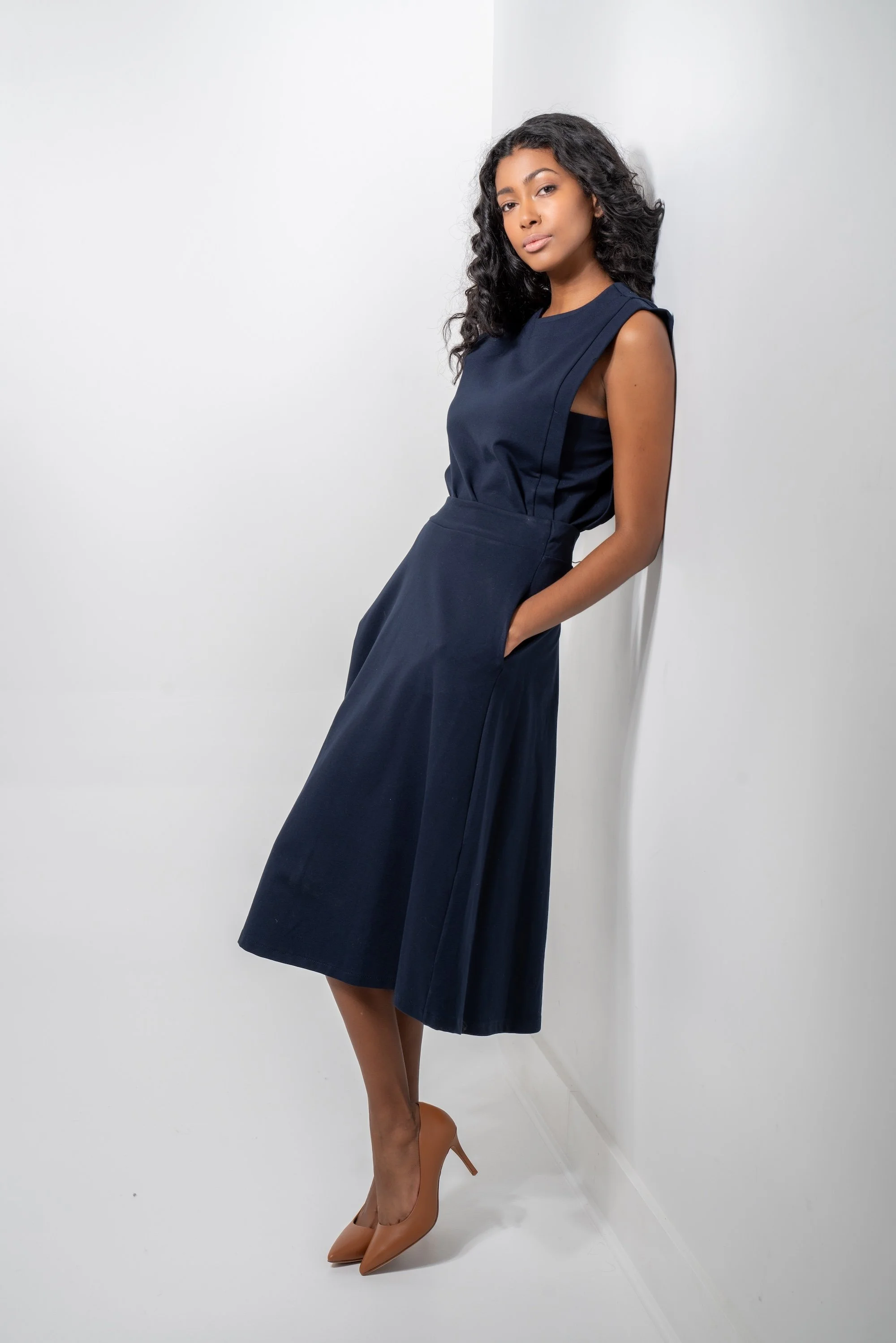 A young woman with curly black hair, wearing a sleeveless navy blue dress and nude high heels, leaning against a white wall with her hands in her pockets.
