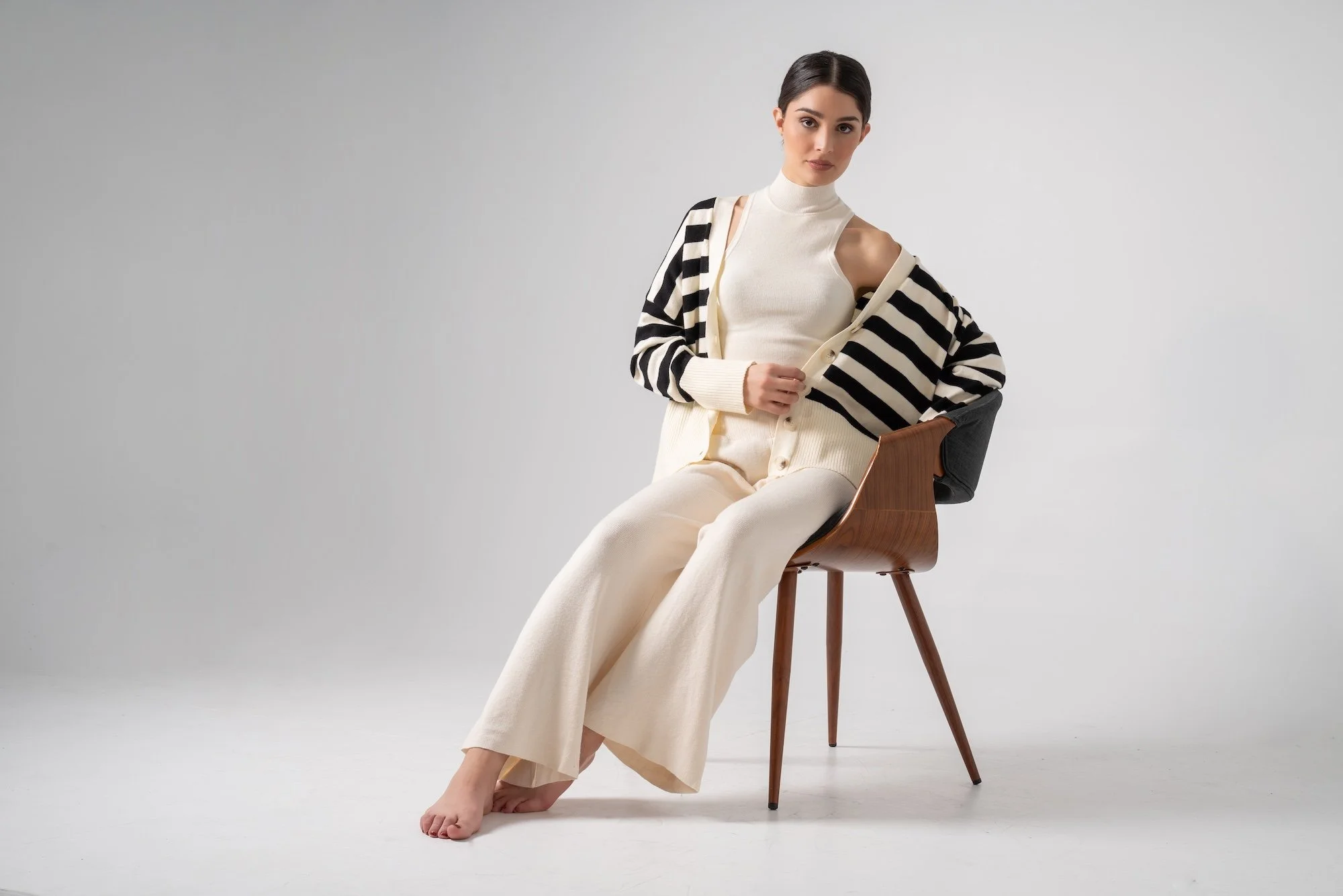 A woman sitting barefoot on a wooden chair, wearing cream-colored pants and a matching sleeveless top, with a black and cream striped cardigan draped over her shoulders, against a plain light gray background.