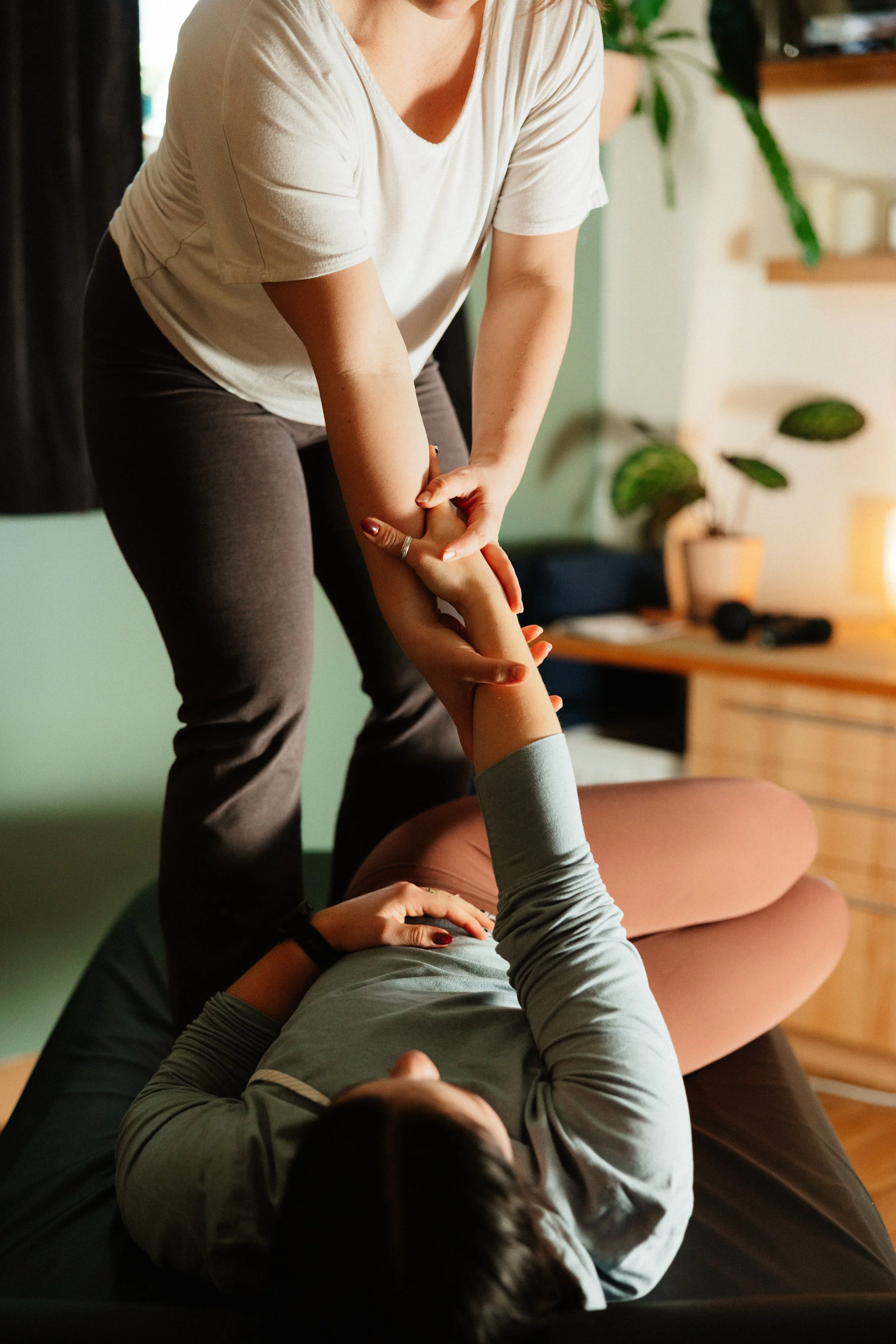 A woman is receiving physical therapy or chiropractic care indoors, lying on a treatment table, while a practitioner holds her arm and elbow.