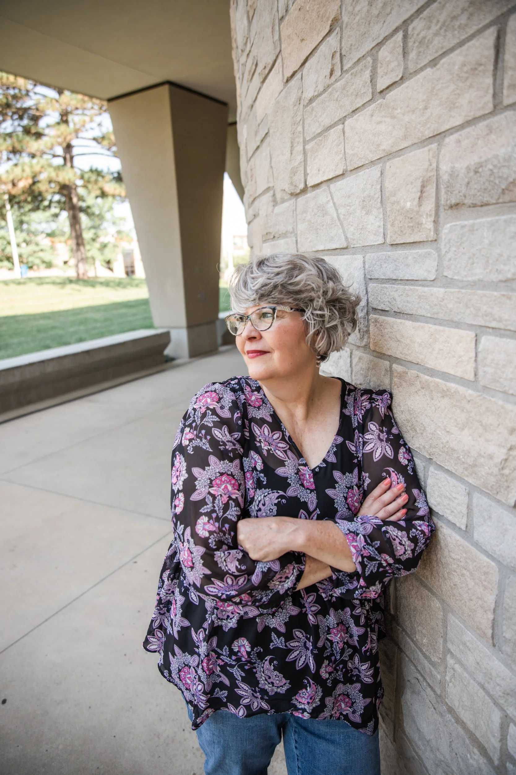 A woman with gray hair and glasses, wearing a black blouse with pink and purple floral patterns, leaning against a beige brick wall outside. She has her arms crossed and is looking to her left. The background includes trees and a building.