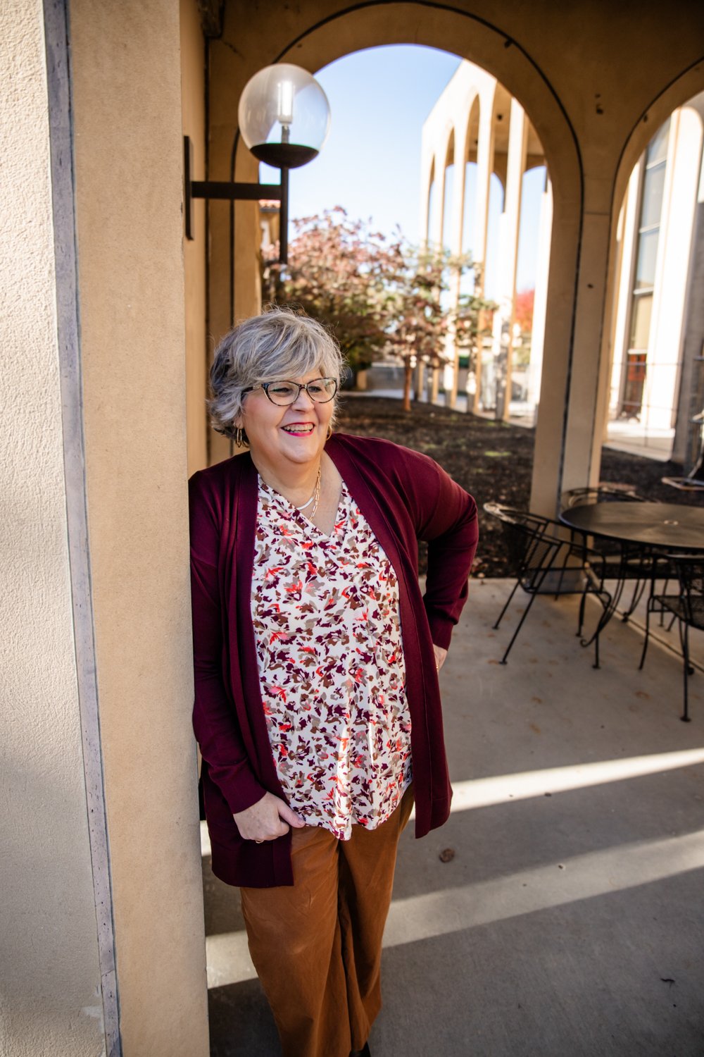 Older woman with gray hair, glasses, and a cheerful smile, leaning against a beige wall outside a building with arched architecture and outdoor seating.