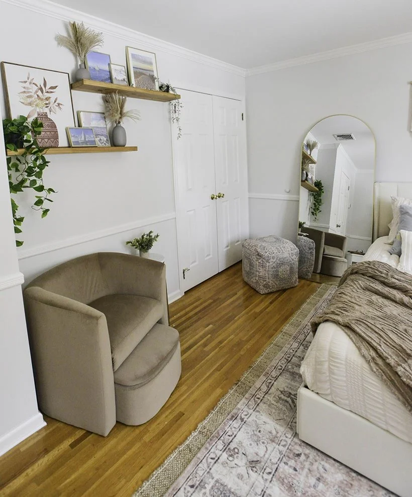 Bedroom corner with beige armchair, wooden shelves with framed art and plants, a large mirror, and a bed with a blanket.