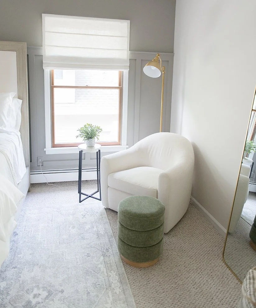 A cozy bedroom corner with a white armchair, a small green ottoman, a side table with a potted plant, a standing lamp, and a mirror reflecting part of the room. There's a window with a white roller shade, and a bed partially visible on the left.