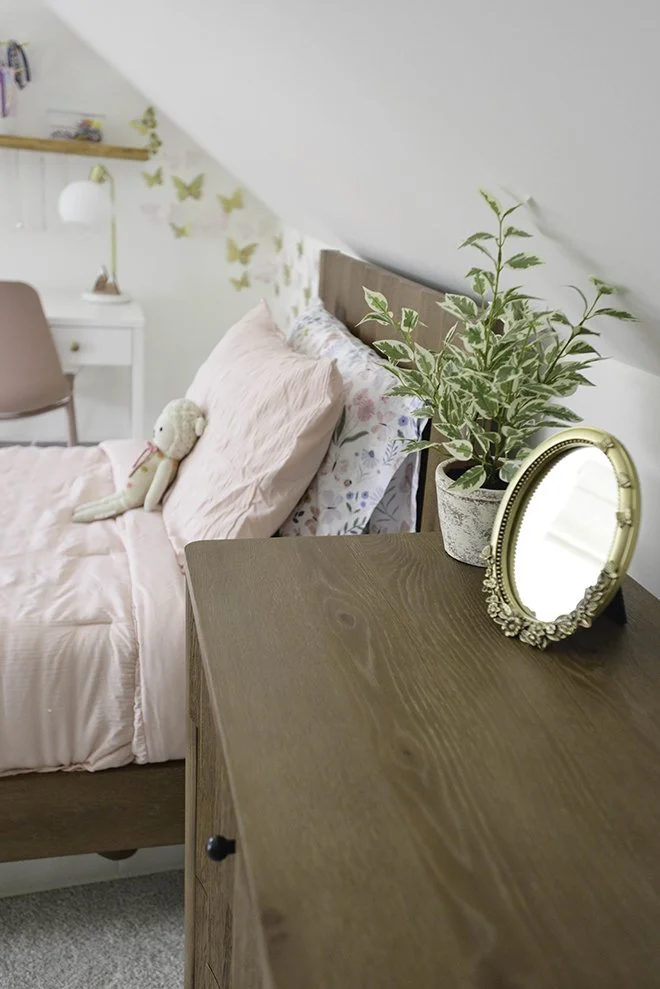 A cozy bedroom with a wooden nightstand holding a potted variegated plant and a decorative oval mirror. The bed has pink bedding and a stuffed doll. In the background, a desk and chair are visible, with butterfly wall decorations.