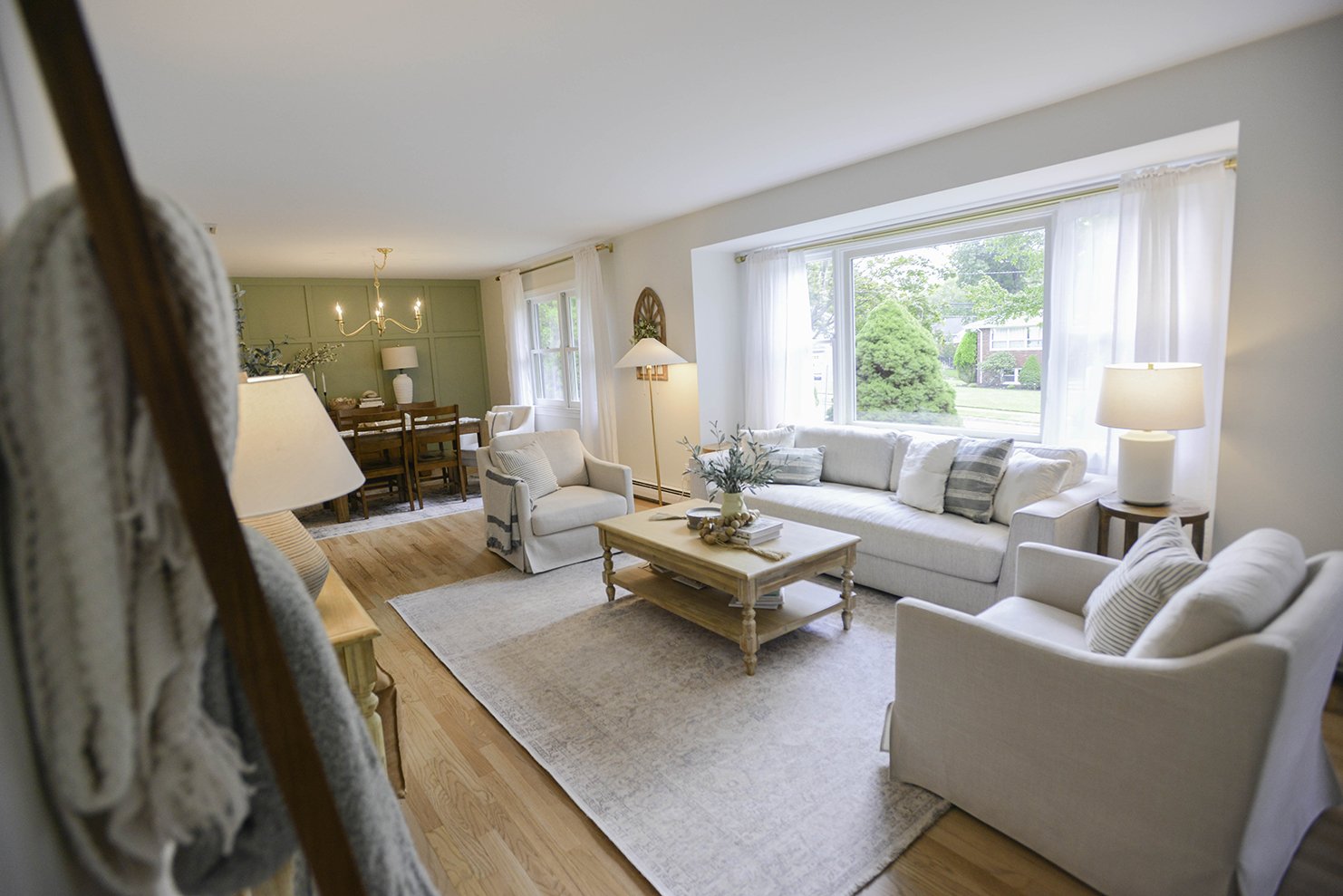 Bright living room with white sofas, wooden coffee table, large window with white curtains, and hardwood floors, view of green backyard outside.