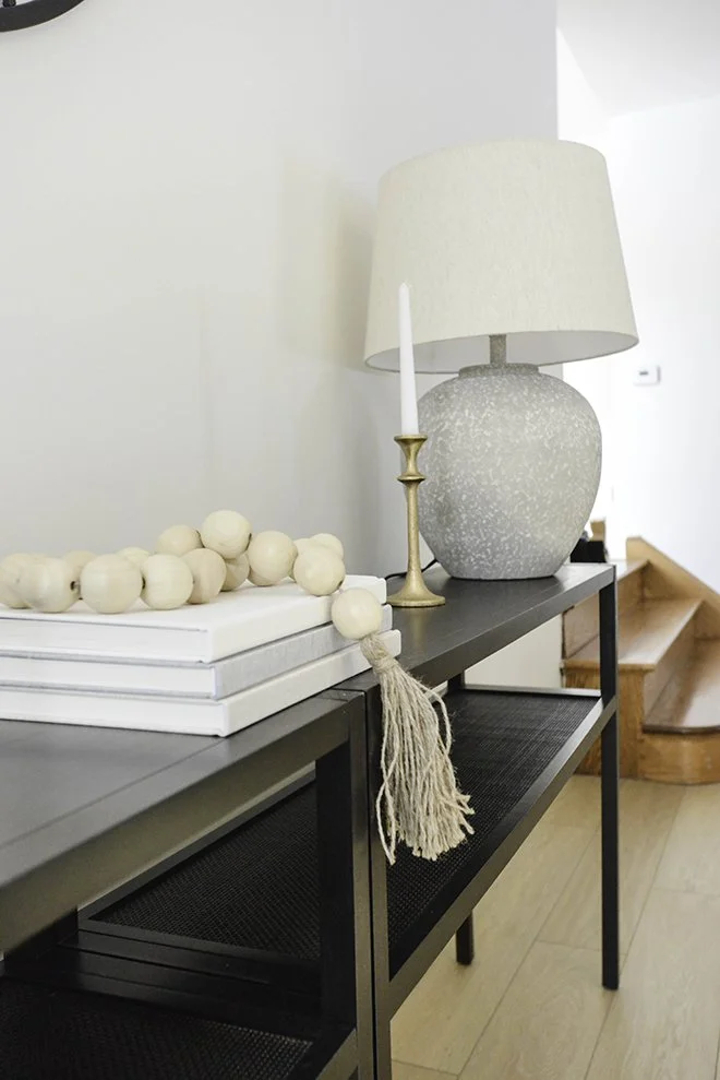 Decorative console table with a large ceramic table lamp, a brass candlestick with a tall white candle, a string of wooden beads, and a stack of three white books inside a modern home with wooden stairs in the background.