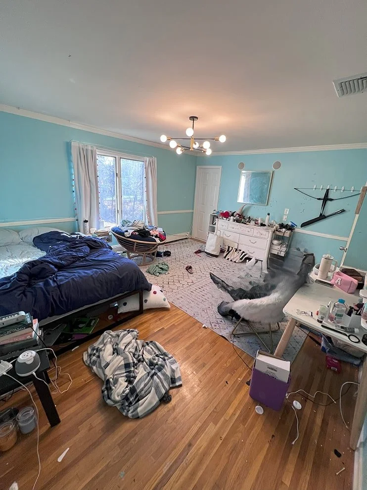 Disorganized bedroom with a spilled blanket on the floor, a large bird pecking at a damp spot on the carpet, a bed with rumpled bedding, and various clutter including clothes, papers, and electronic devices scattered around.