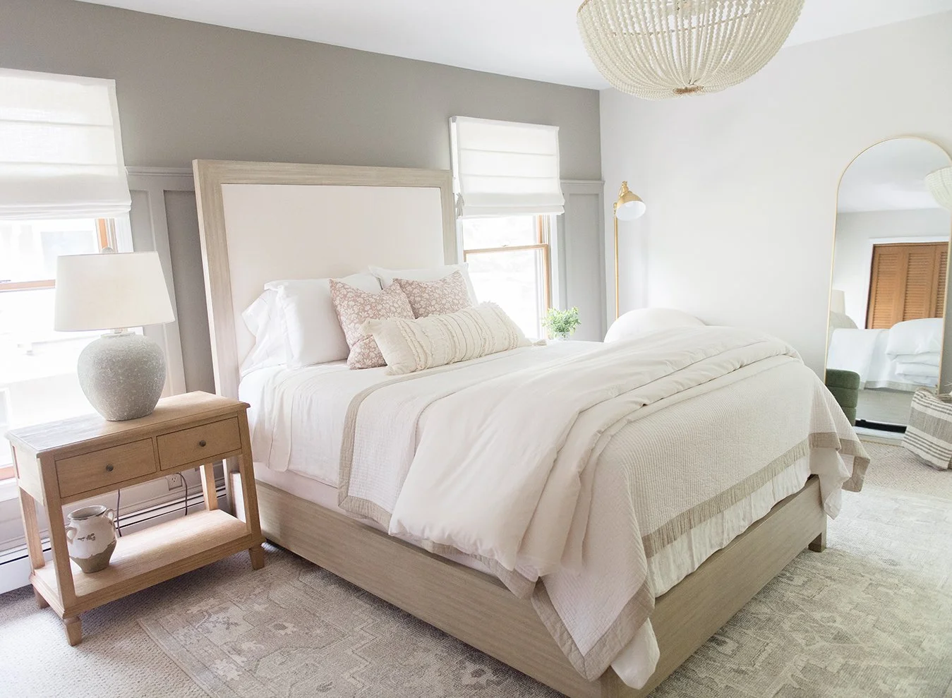 Bright, minimalist bedroom with a large white bed, beige bedding, and decorative pillows. Light wood nightstand with vase and lamp, windows with white blinds, and a mirror reflecting part of the room.