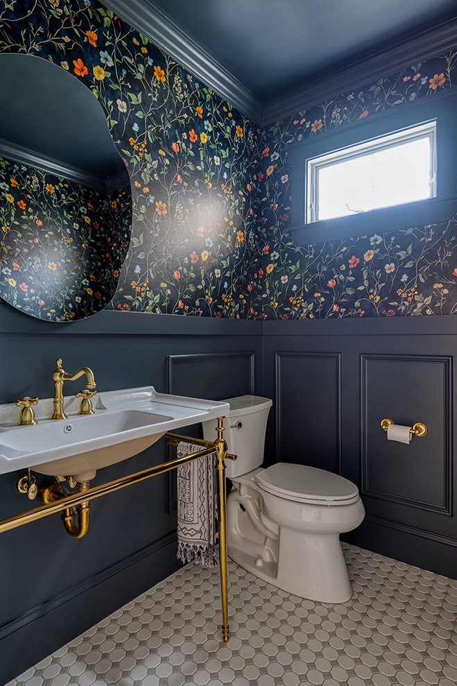 A bathroom with dark floral wallpaper, a large oval mirror, a white porcelain sink with gold fixtures, a white toilet, a gold toilet paper holder, and a small window letting in natural light.