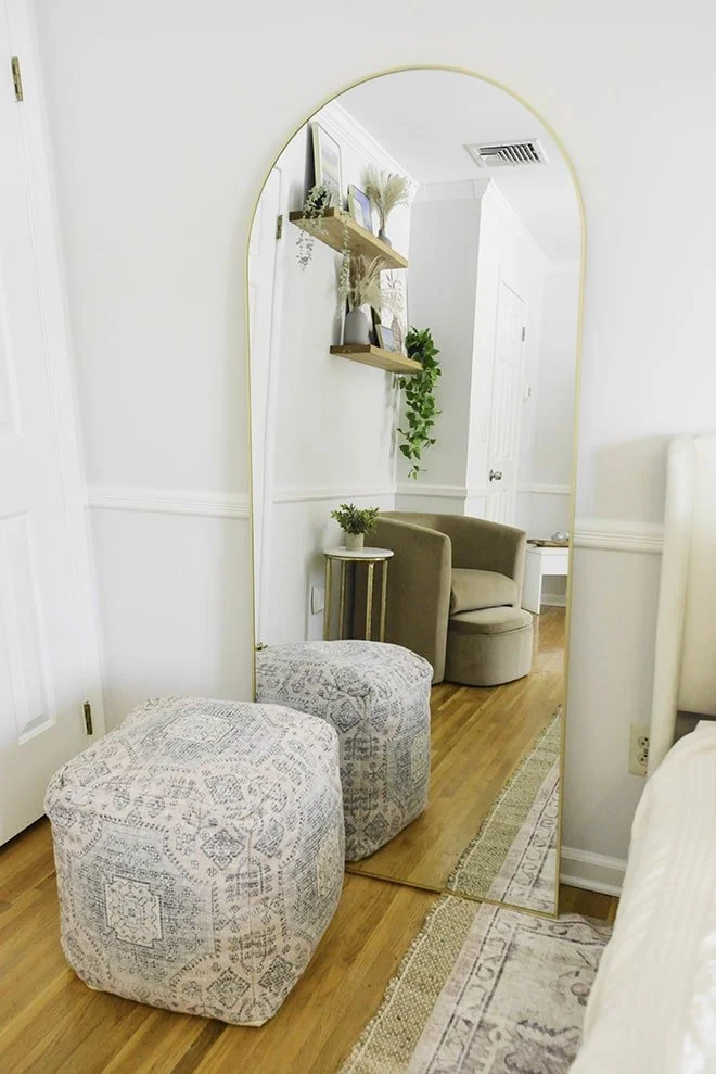 A tall, arched mirror reflecting a cozy corner of a room with beige armchair, small side table with a plant, two floating shelves with decorative items, and a wooden floor with a rug.