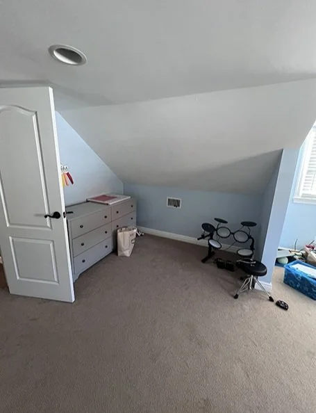 Empty room with sloped ceiling, gray dresser, small black drum set, blue storage bin, and window with blinds.