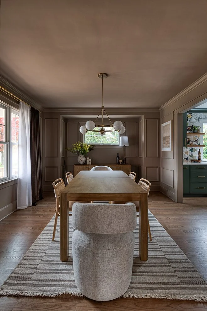 Modern dining room with a wooden table, six rattan chairs, a striped area rug, a large window with white curtains, and a chandelier with globe lights.
