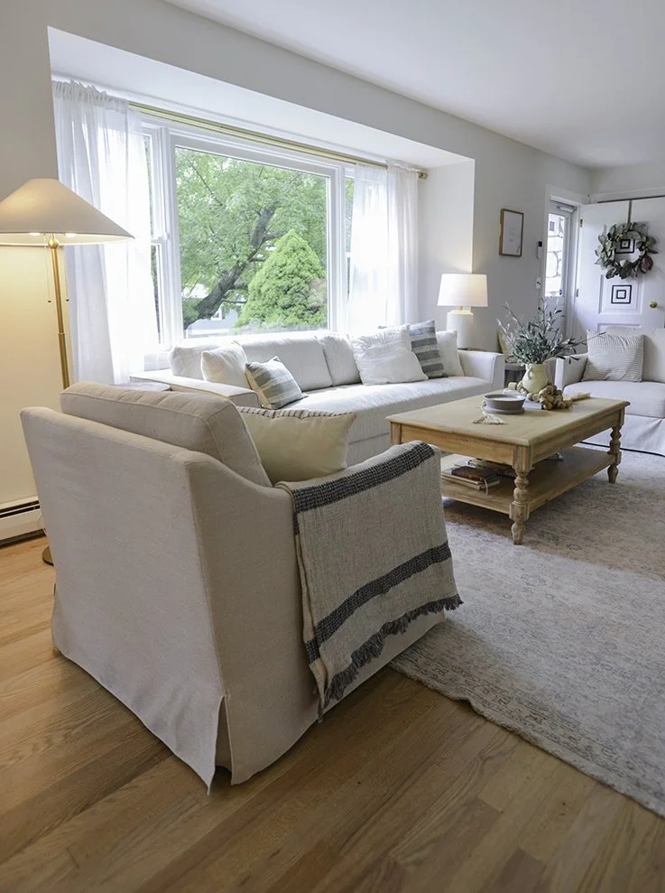 A cozy living room with a large window, white curtains, white sofas with pillows, a wooden coffee table with decorative items, a floor lamp, and a door with a wreath, all in a bright, airy space with wooden flooring.