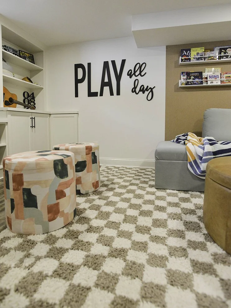 A cozy living room with a patterned carpet, a gray sofa with a striped blanket, wall shelves with books, and a large wall decal saying 'PLAY all day'.