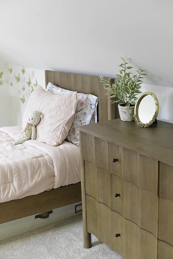 A cozy bedroom corner with a wooden bed, pink bedding, a stuffed animal, and a dresser holding a plant and a round mirror.