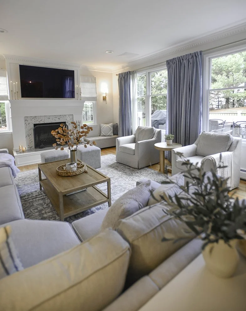 Bright living room with white and beige furniture, a wooden coffee table with decorative vase and flowers, fireplace with tile surround, large windows with blue curtains, and a view of a patio outdoors.