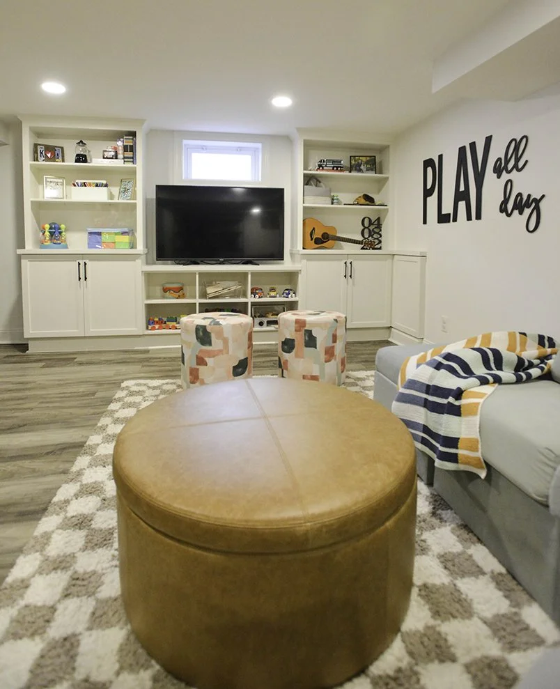 A cozy living room with built-in shelves, a large TV, a small window above the TV, an orange guitar, and a wall sign that says 'PLAY all day.' There is a coffee table, a patterned ottoman, a gray sofa with a striped blanket, and a patterned rug.
