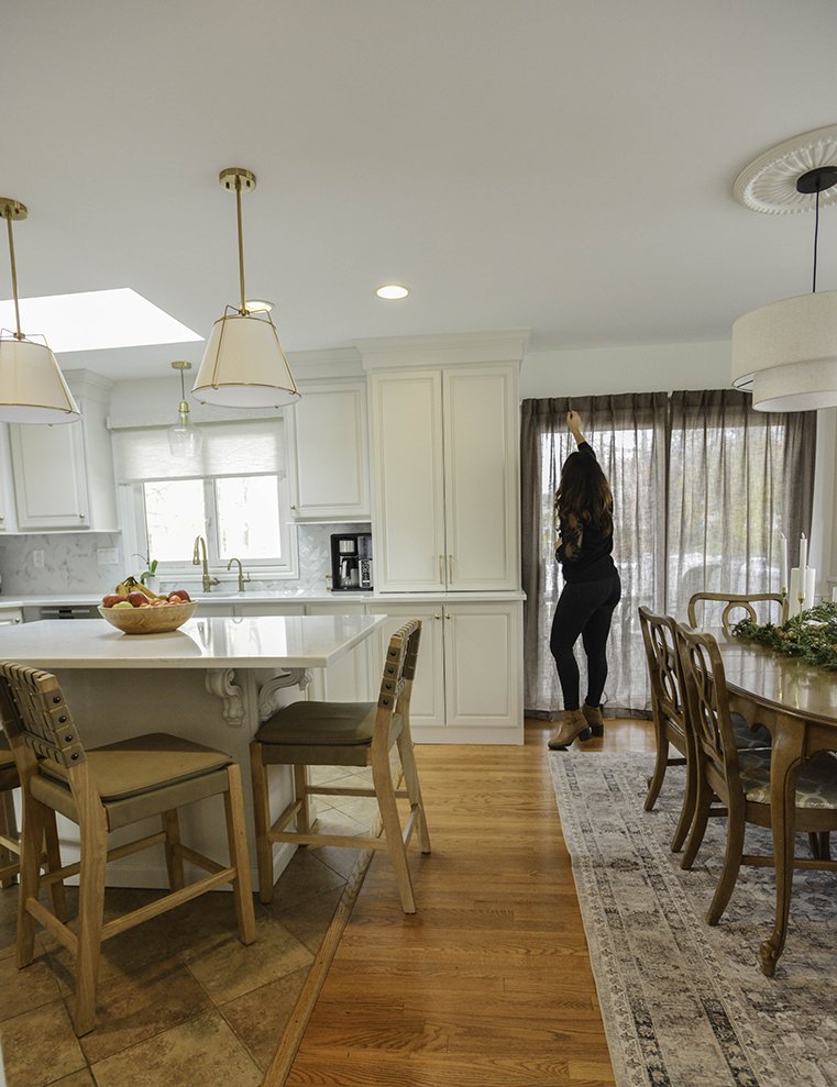 A woman with long brown hair, dressed in black, standing in a bright, modern kitchen with white cabinets, wood flooring, and a dining area, reaching up towards the ceiling curtain.