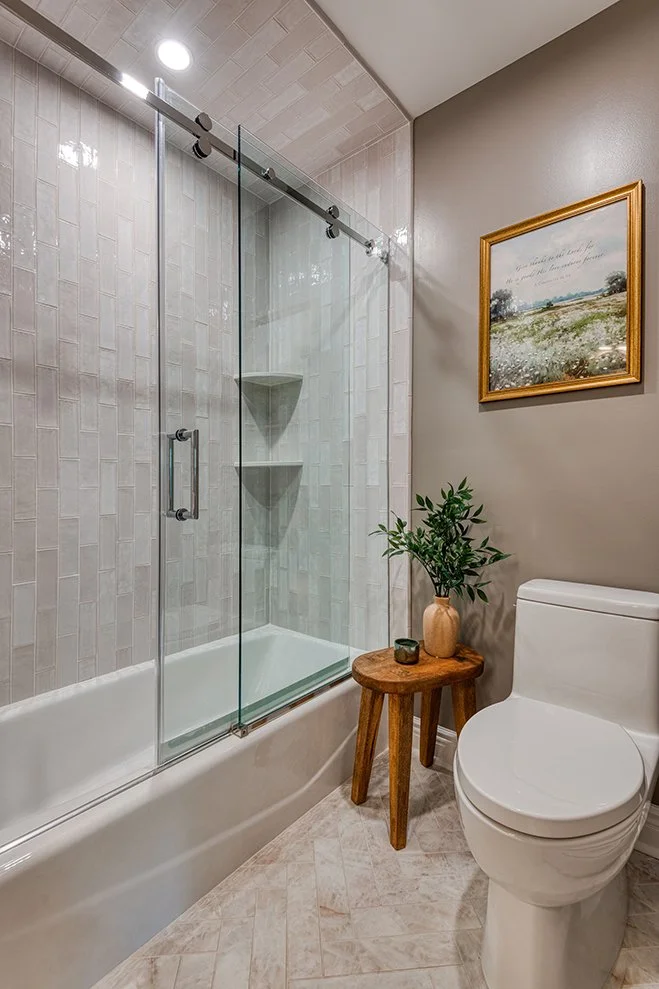 A modern bathroom with a glass-enclosed shower, a white toilet, a small wooden side table with a potted plant, a ceramic vase, and a framed landscape painting on the wall.