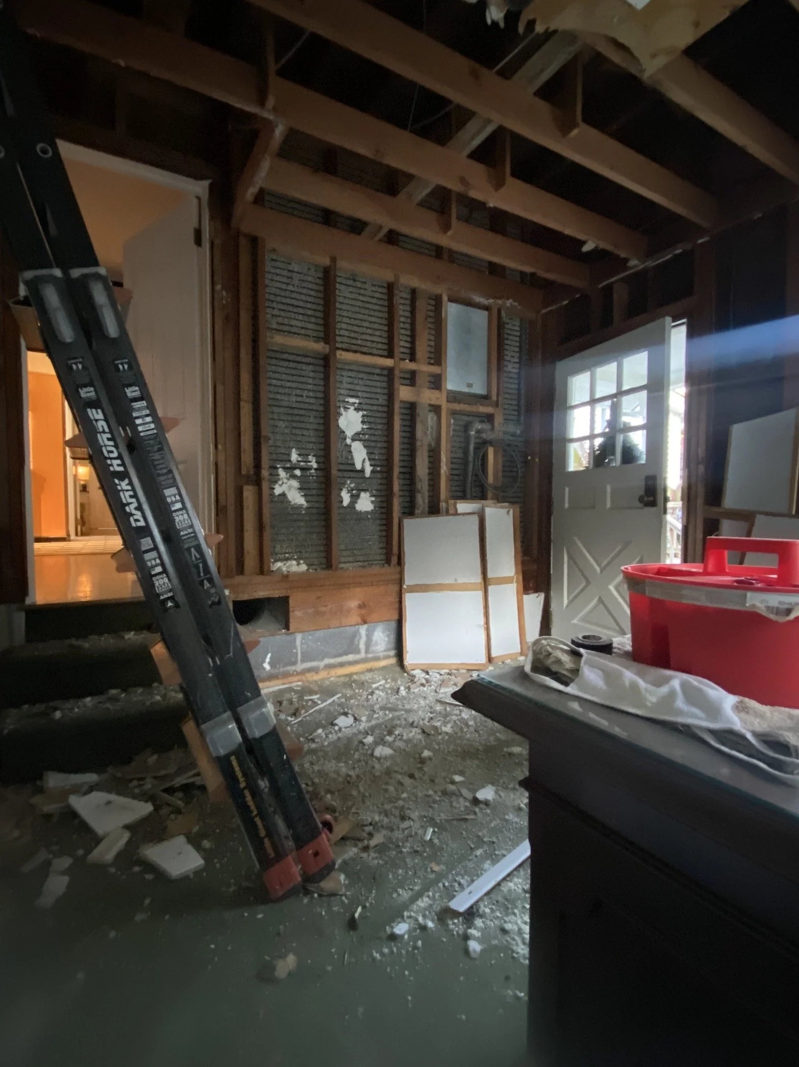 Interior of a room under renovation with exposed wooden framing, a door with glass, a red container, a cloth on a black piece of furniture, a construction level leaning against the wall, construction debris on the floor, and daylight coming through t