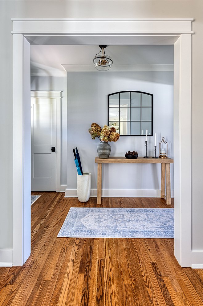 View through a white doorway into a hallway with a wooden floor, a small wooden console table holding decor items, a large wall mirror, and a vase with dried flowers.