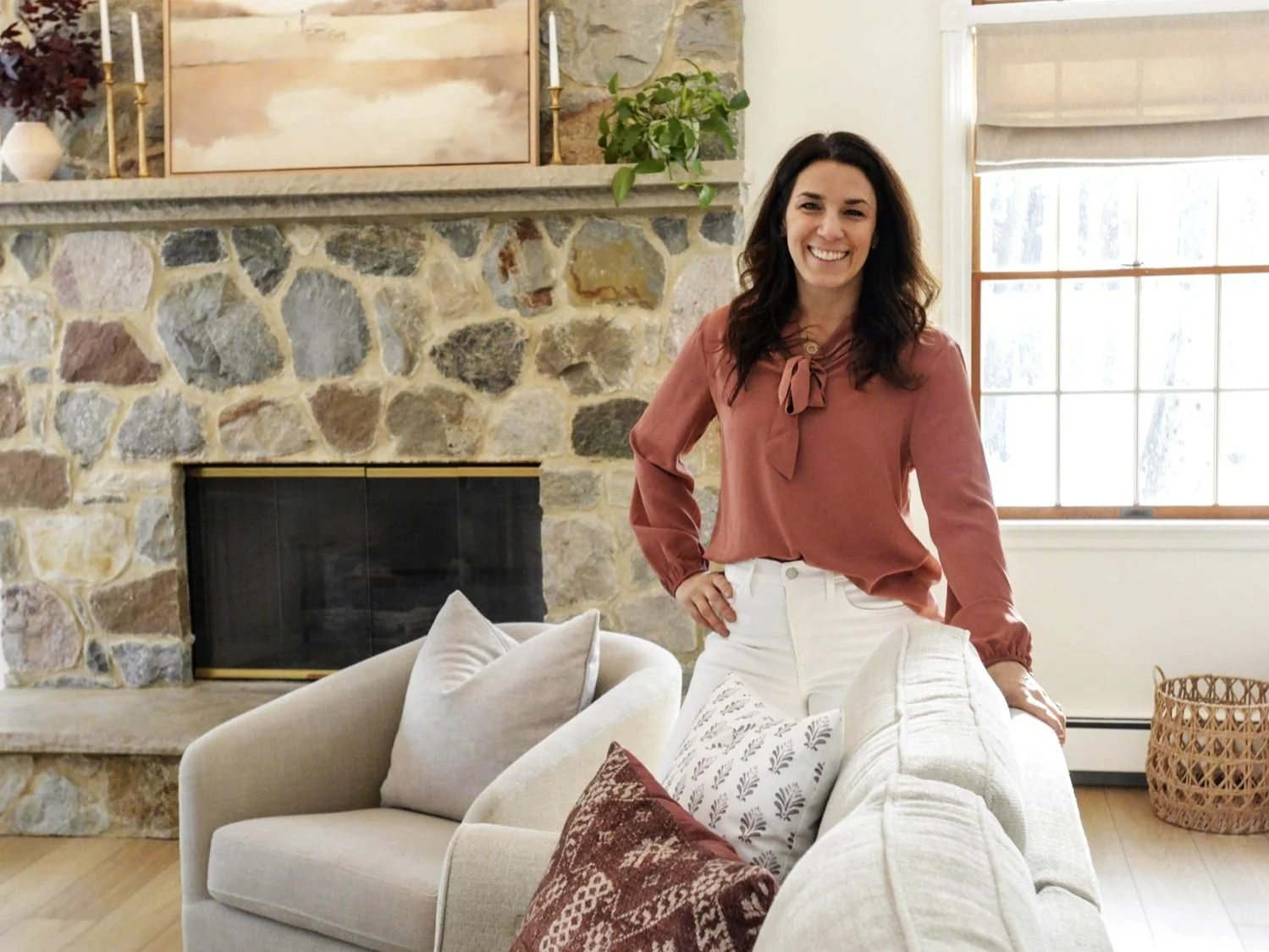 A woman with dark hair wearing a pink blouse and white pants, smiling and standing behind a beige sofa with decorative pillows. The background features a stone fireplace, a painting, and a window with a view of snow outside.