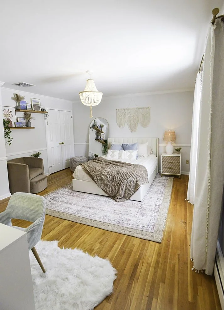 A cozy bedroom with white walls, hardwood floors, a large bed with white and gray bedding, a chandelier, and a window with curtains. There are decorative items like plants, framed pictures, a macramé wall hanging, and a nightstand with a lamp.