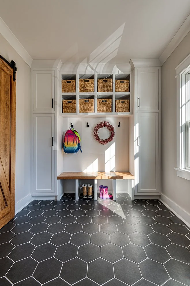 Entryway with white cabinets, a wooden bench, and wicker baskets above it, a colorful backpack hanging, pink rain boots, and a pink wreath on the wall, with hexagonal black floor tiles and sunlight coming through a window.