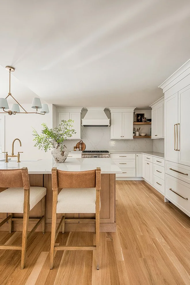 Modern kitchen with white cabinets, wooden flooring, and a central island with a vase of greenery. There are two beige chairs at the island and a brass chandelier overhead.