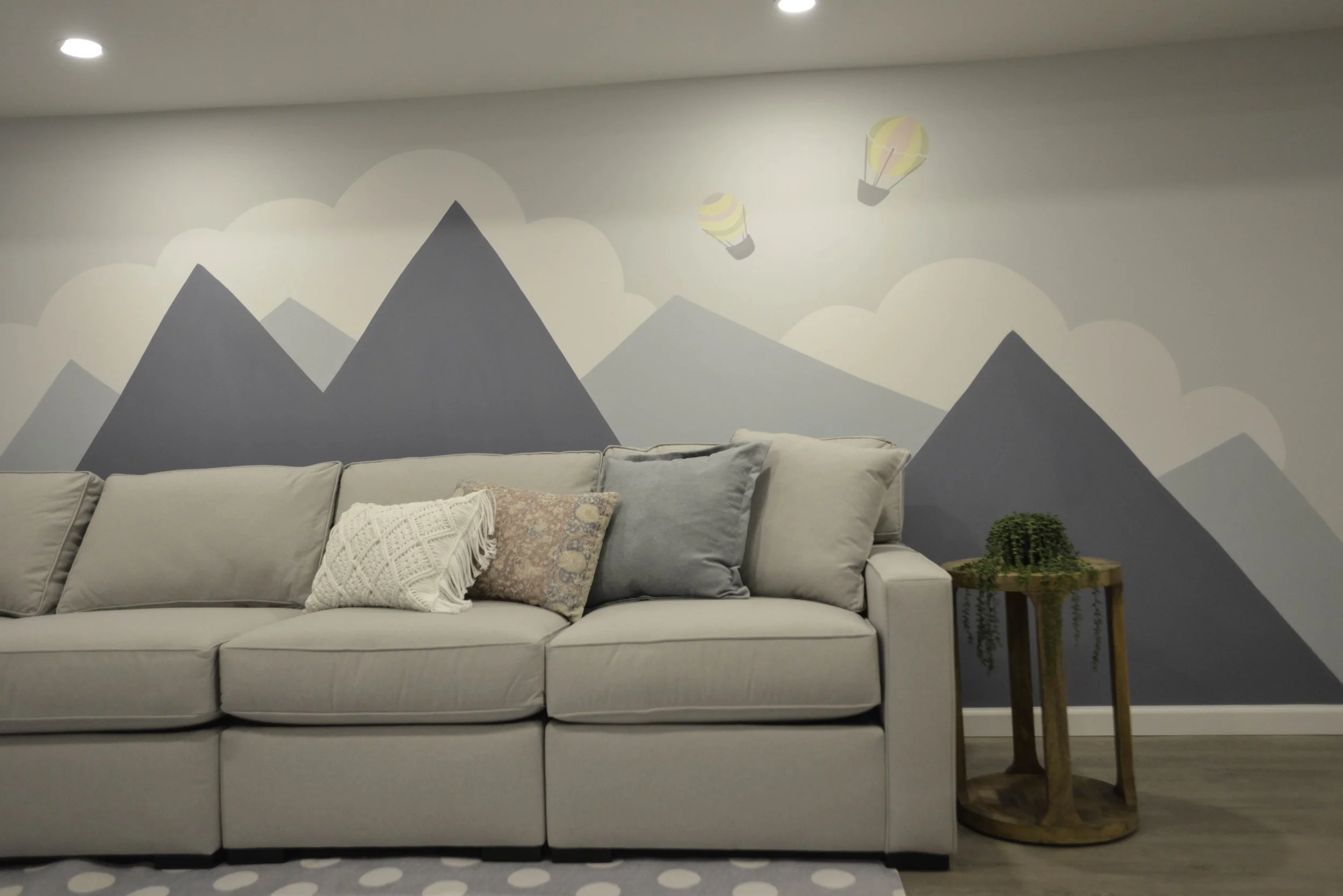 Living room with a beige couch and colorful pillows, wall mural of mountains and hot air balloons, wooden side table with a green fern plant.