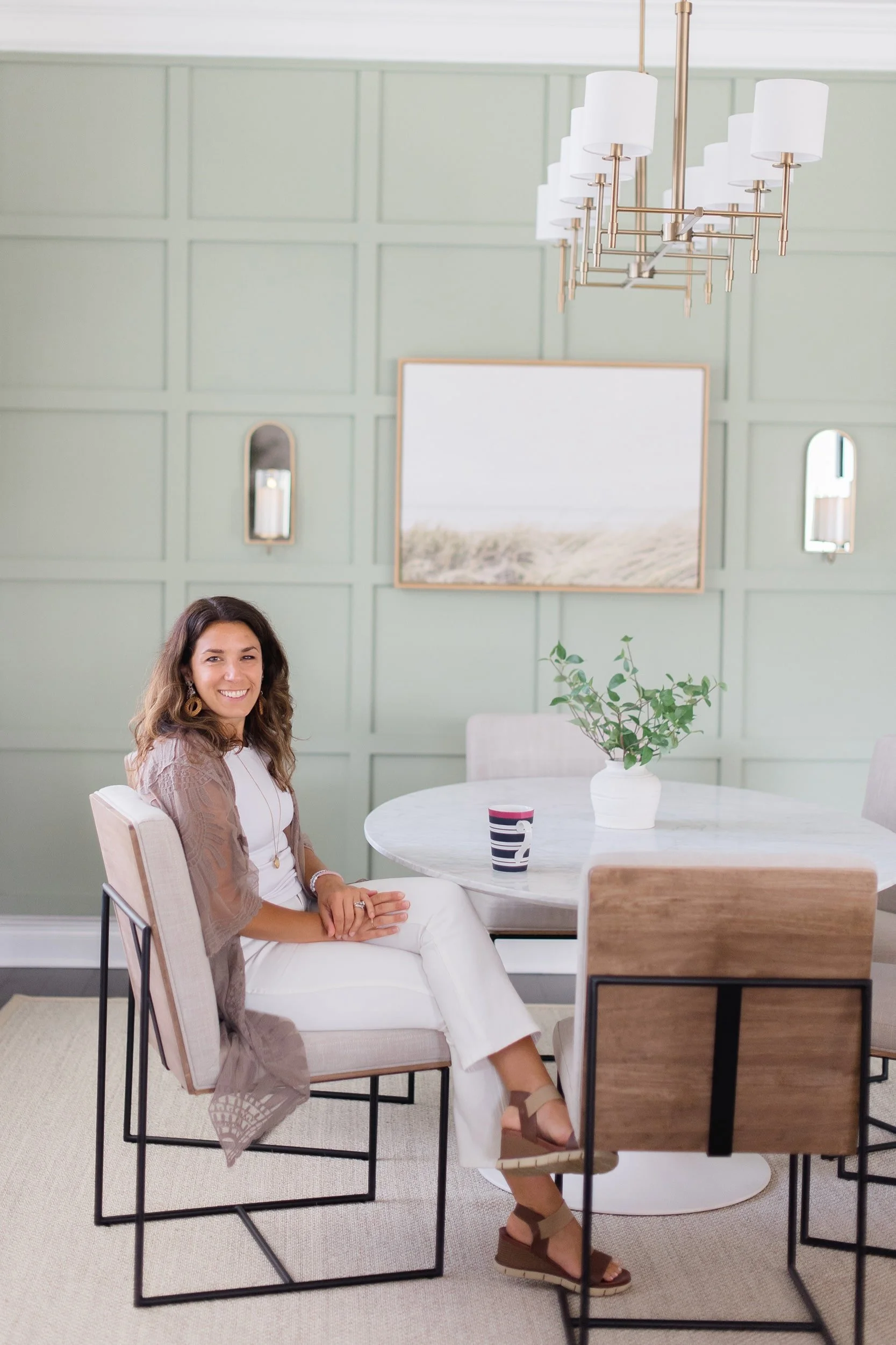 A woman sitting at a round dining table in a modern, light green-walled room with a chandelier, a framed landscape picture, and two small wall mirrors.
