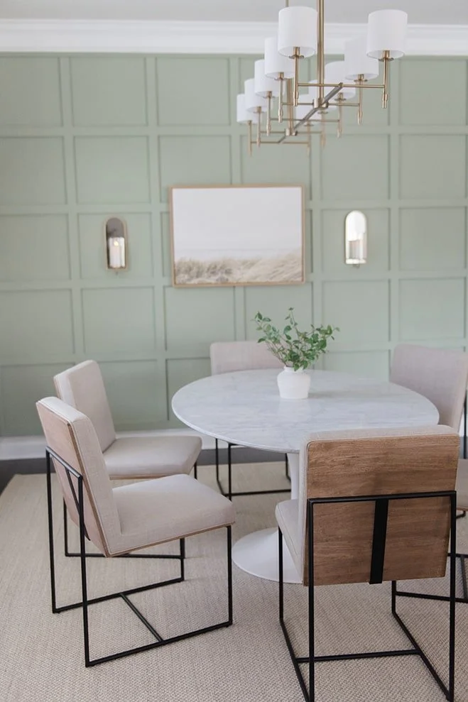 Modern dining room with a round marble table, four chairs with cushioned seats and backs, and a branch of green plant in a white pot on the table. Light green paneled wall with framed art and two wall sconces, large white and gold chandelier overhead