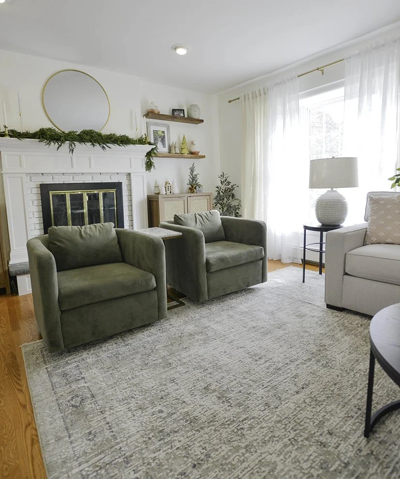 A cozy living room with two green armchairs and a white sofa around a gray area rug. There is a white brick fireplace with a mirror above, decorated for Christmas with greenery. The room has large windows with white curtains and various decorative it