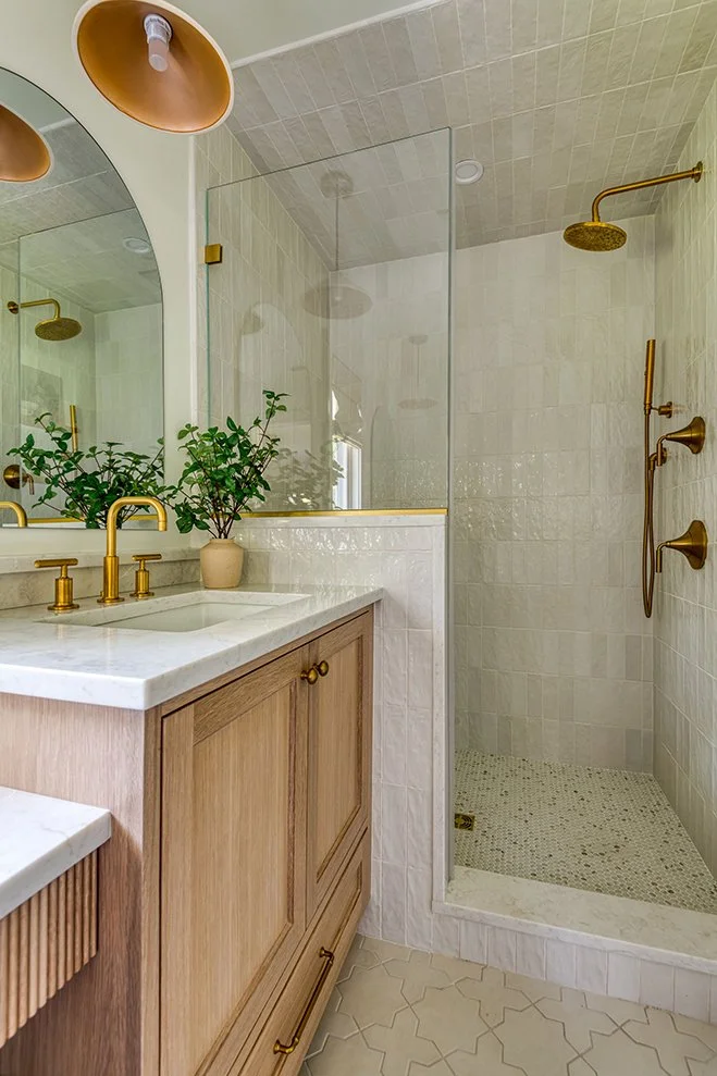 Modern bathroom with a wooden vanity, white marble countertop, and brass fixtures, including a faucet and showerhead, with a potted plant on the counter and a large mirror above the vanity. The walk-in shower has a glass partition and beige tile wall