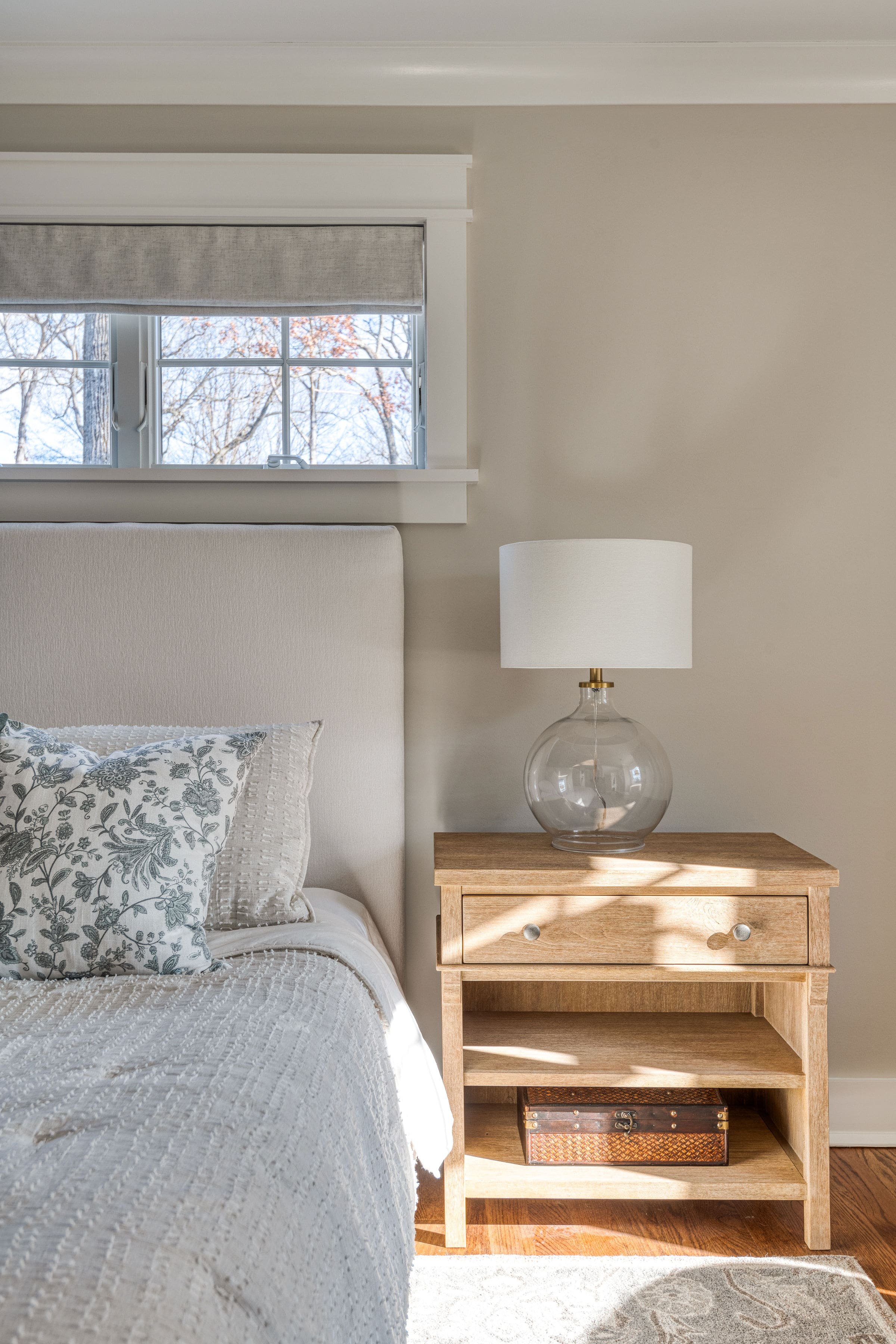 A bedroom with a beige upholstered bed, pillows with floral and textured patterns, a wooden bedside table with a glass lamp, and a window with a view of trees outside.