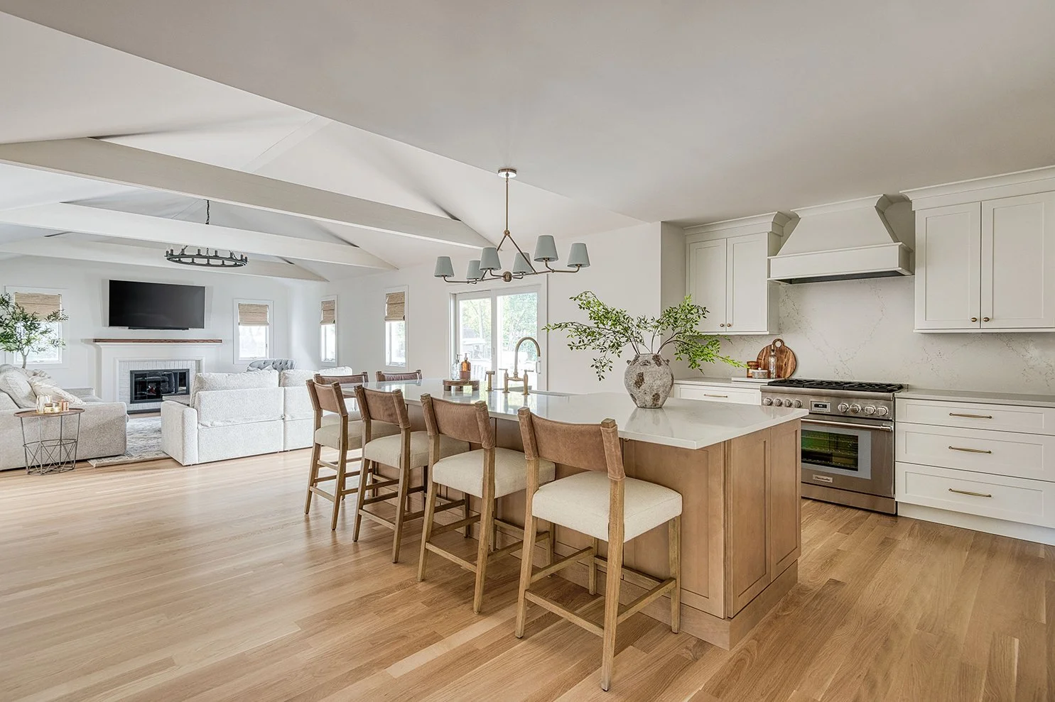 Open-concept kitchen and living room with hardwood floors, white cabinetry, modern appliances, a large island with bar stools, a living area with sofas, a wall-mounted TV, and a fireplace.