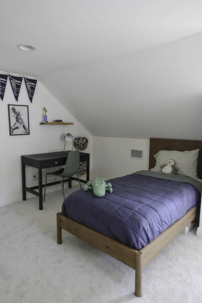 A cozy bedroom with a sloped ceiling, a bed with a wooden frame, dark blue quilt, and stuffed animals. A small desk with a gray chair is against the wall, with a framed picture and a shelf above. The room has light-colored carpet and white walls.