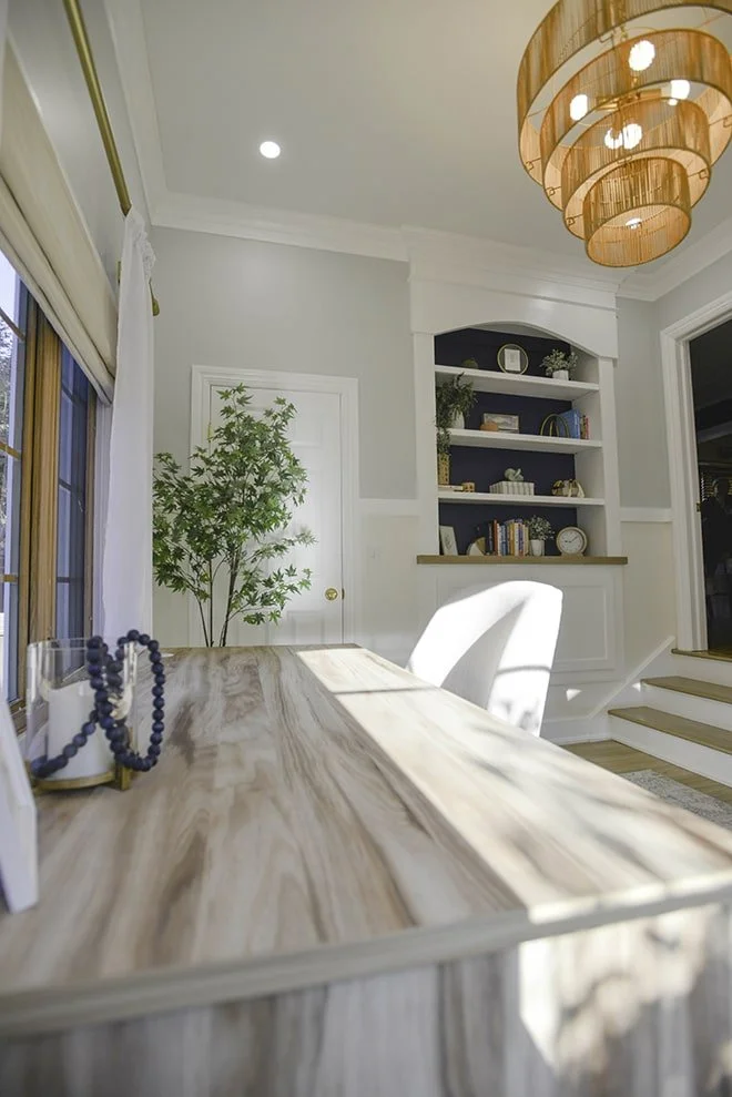 Bright home office with a wooden desk, white chair, potted plant, built-in bookshelf, and modern chandelier.