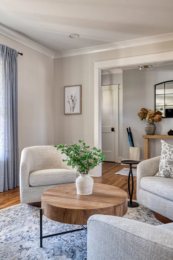 A cozy living room with beige upholstered armchairs, a round wooden coffee table with a green potted plant, a patterned rug, and a light-colored wall with a framed artwork. There's a window with blue curtains and a doorway leading to another room wit
